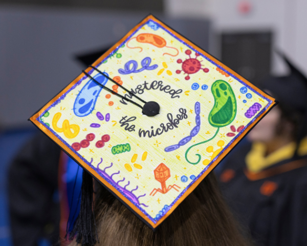 Photo of a graduate wearing an academic cap that is decorated with illustrations of microorganisms and text that reads, 